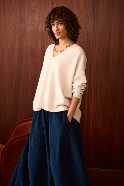 A person with curly hair stands in front of a wood-paneled wall, wearing a cream sweater and the Jumbo Cord Bubble Skirt in blue, with hands in pockets.