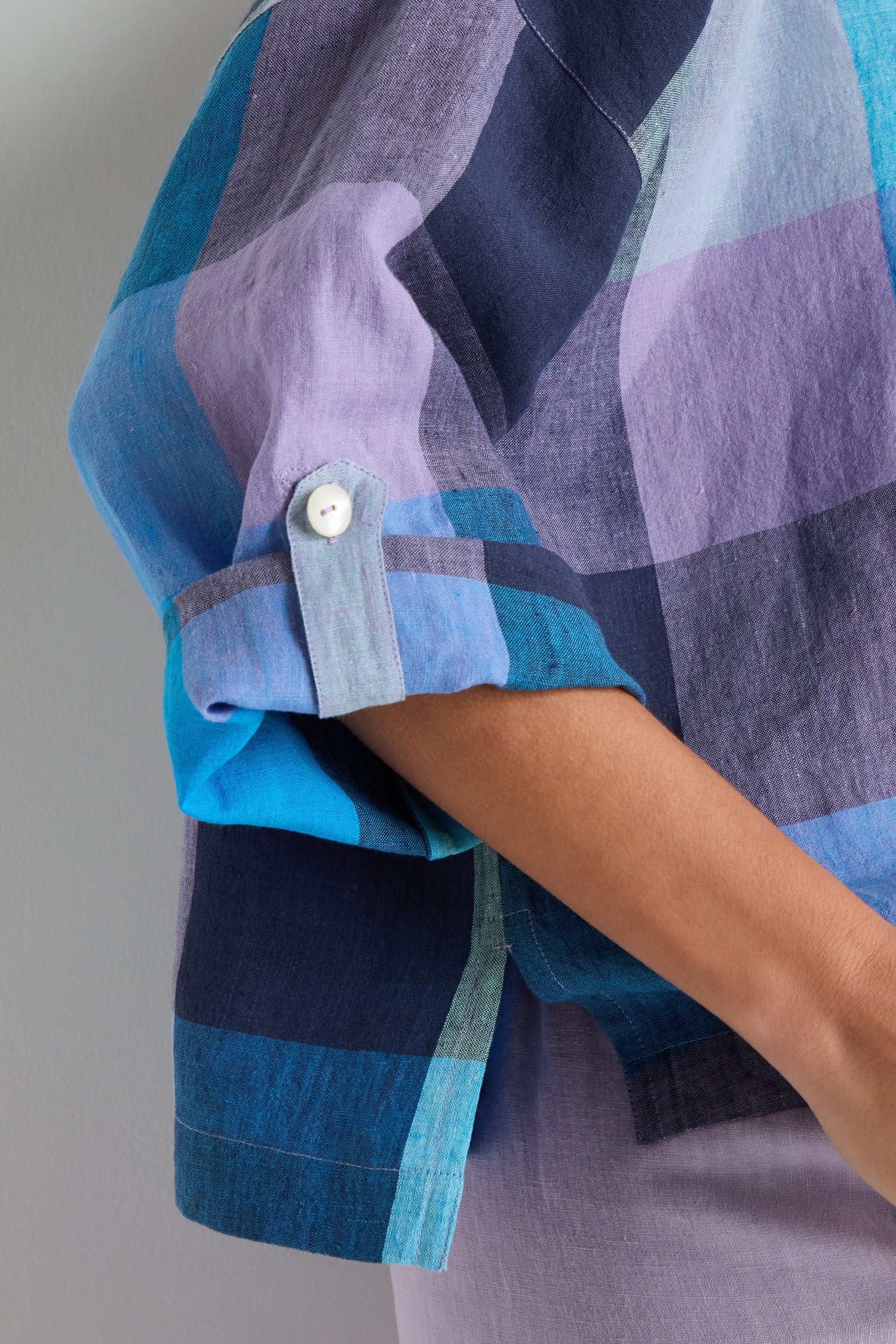 Someone wears the Cross Dye Check Linen Shirt, a short-sleeved, yarn-dyed linen shirt with an oversized fit and purple, blue, and dark blue plaid. Rolled-up sleeves fasten with buttons for a relaxed, casual style.