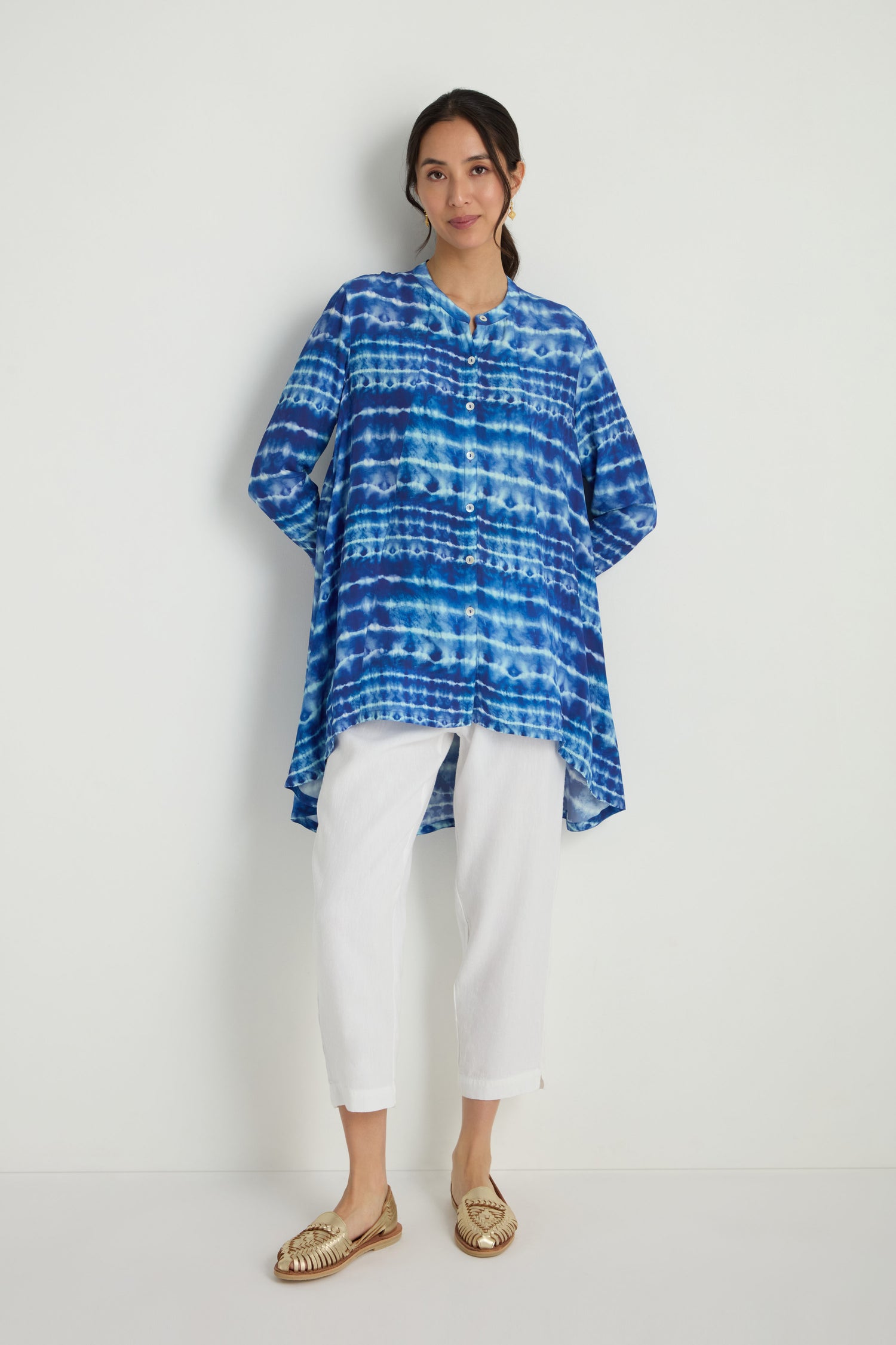 A woman stands against a white wall wearing a Tie Dye Printed Viscose Shirt, white pants, and gold woven shoes.