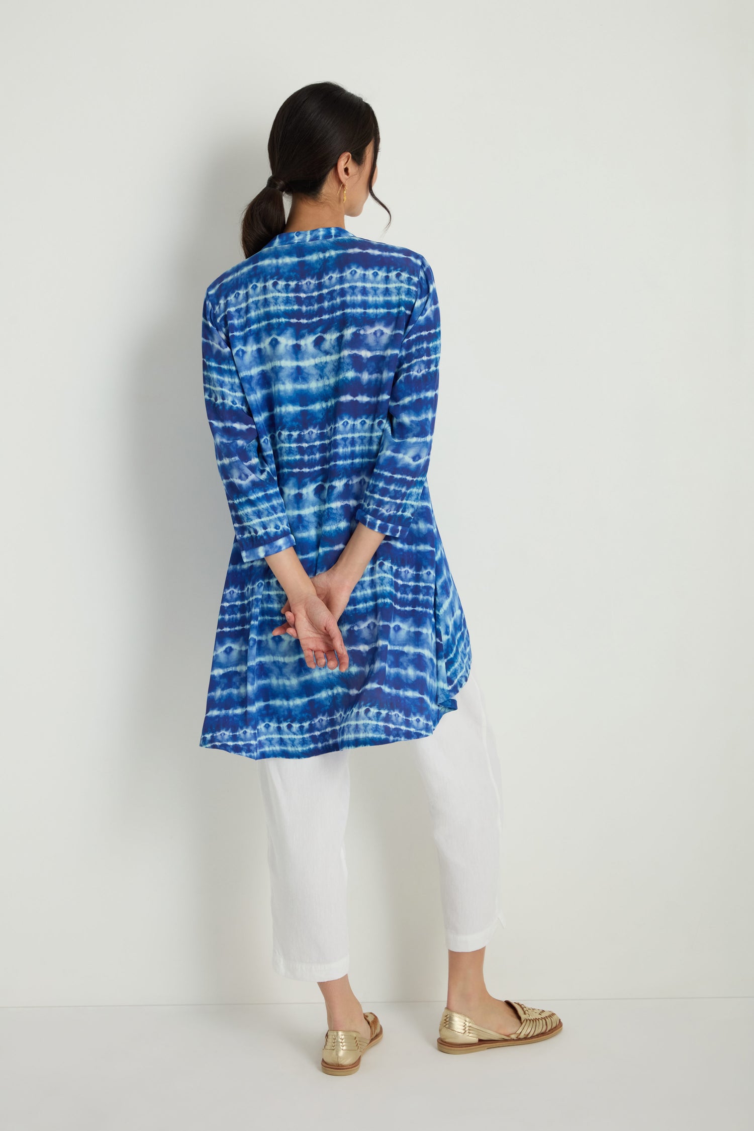 A woman faces a white wall, wearing the Tie Dye Printed Viscose Shirt styled as a tunic with white pants and gold shoes, her hands clasped behind her back.