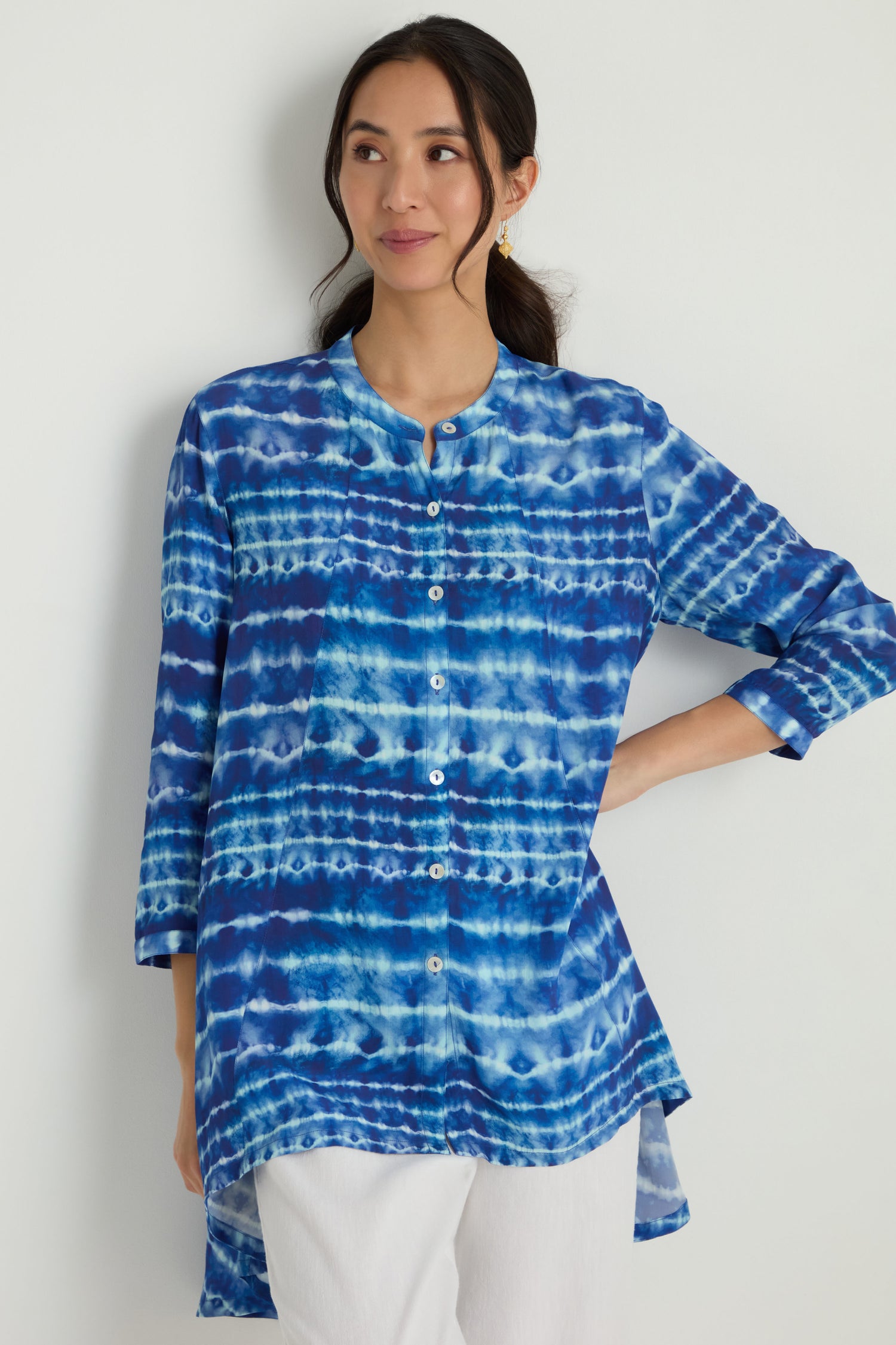 A woman stands against a white wall wearing the Tie Dye Printed Viscose Shirt styled as a blue and white button-up tunic with three-quarter sleeves, paired with white pants.