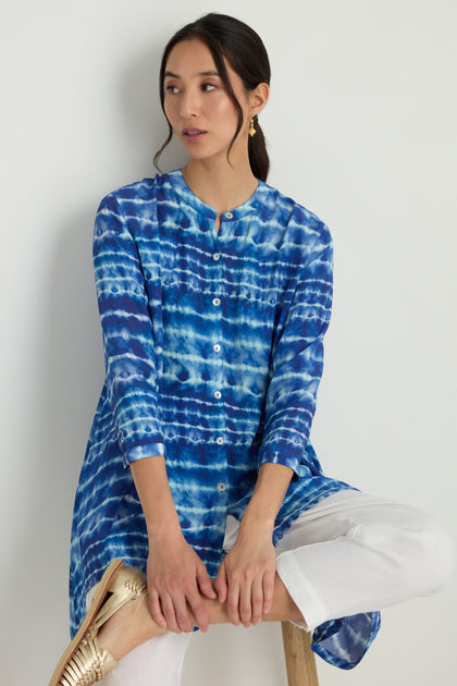 A woman sits on a stool, wearing the Tie Dye Printed Viscose Shirt paired with white pants and gold flats, looking to her left against a plain light background.