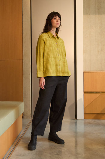 A woman stands indoors wearing the Brushed Cotton Pintuck Detail Shirt in yellow, paired with loose black pants and black shoes, with neutral-toned walls and furniture in the background.