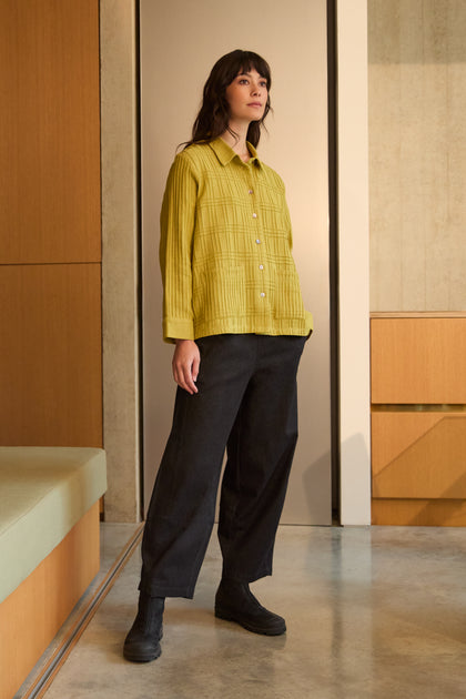 A woman stands indoors wearing the Brushed Cotton Pintuck Detail Shirt in yellow, paired with loose black pants and black shoes, with neutral-toned walls and furniture in the background.