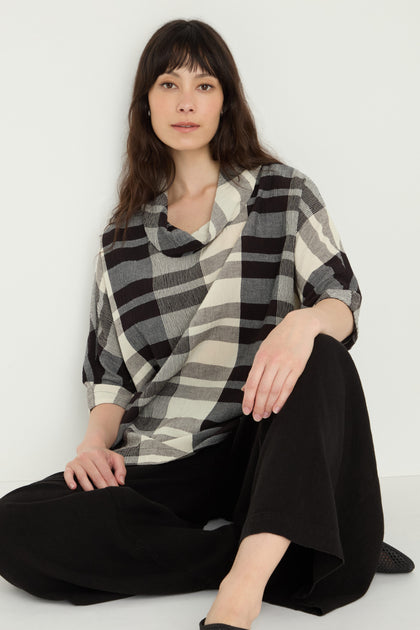 A person with long brown hair, wearing a Monochrome Crinkle Check Cowl Neck Top and black wide-leg pants, sits on the floor against a white wall.