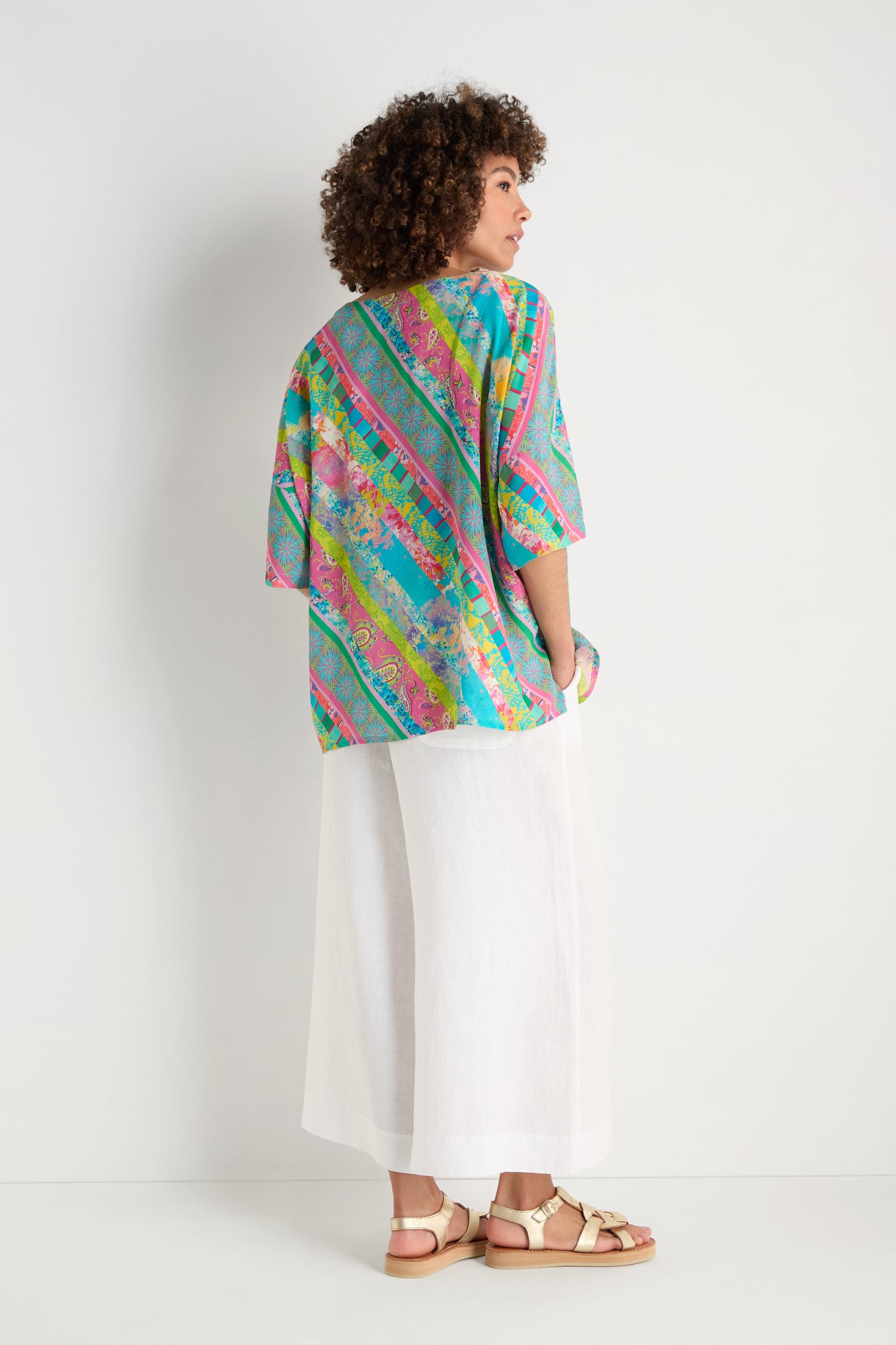A person stands sideways in a Patchwork Stripe Boxy Top, wide white pants, and beige sandals against a plain white background.