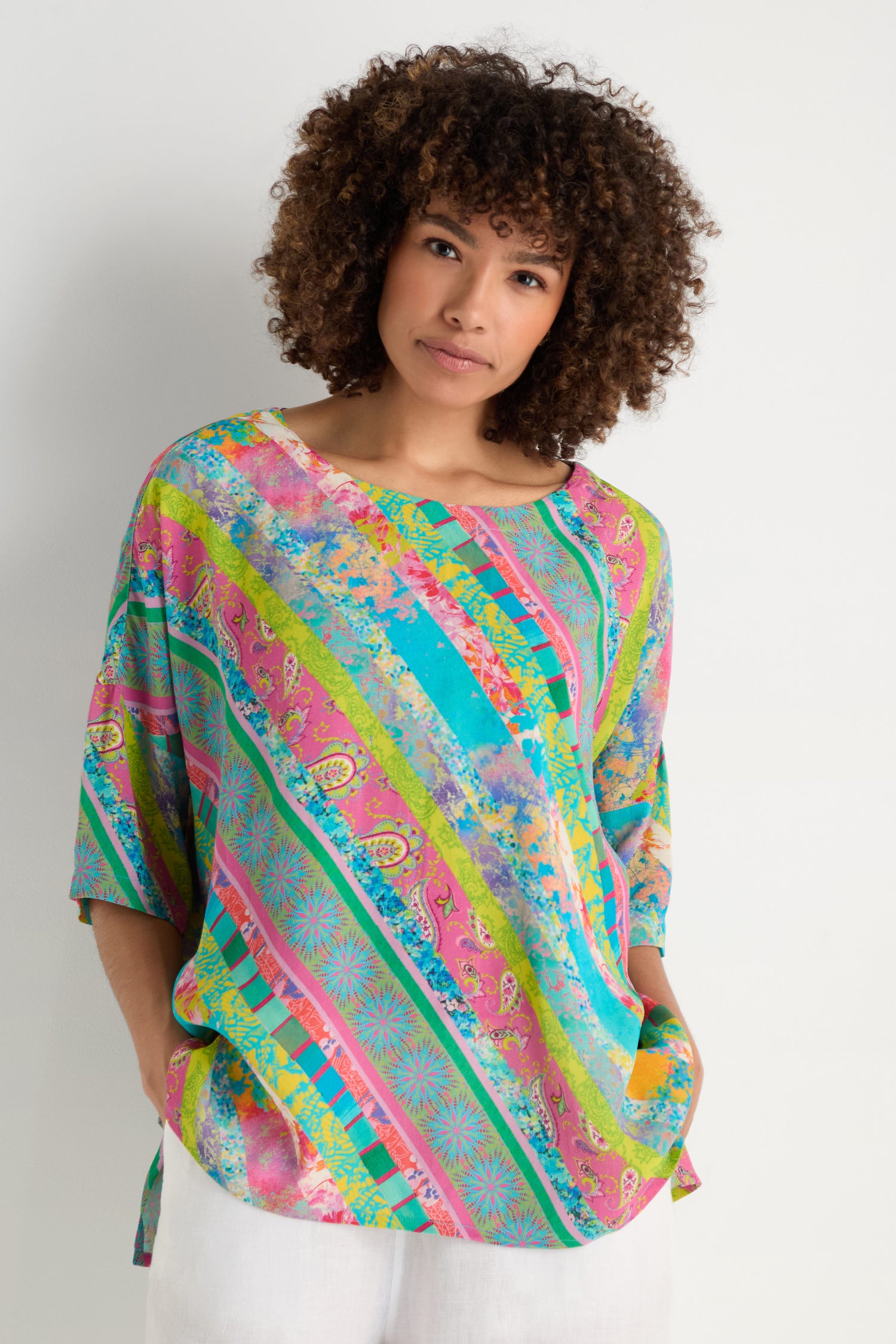A woman with curly hair wears the Patchwork Stripe Boxy Top in pink, blue, and green archive prints, paired with white pants, standing against a plain white background.