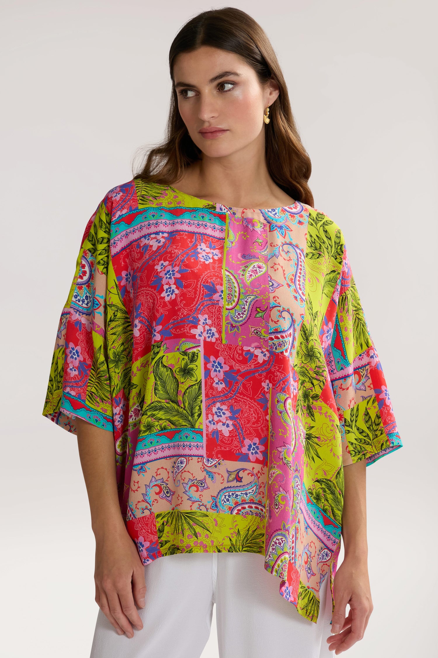 A woman wearing a Tropical Patchwork Relaxed Top made of pure viscose, with a bohemian style featuring red, pink, green, and blue patterns, stands against a plain background. She is looking slightly to the side.