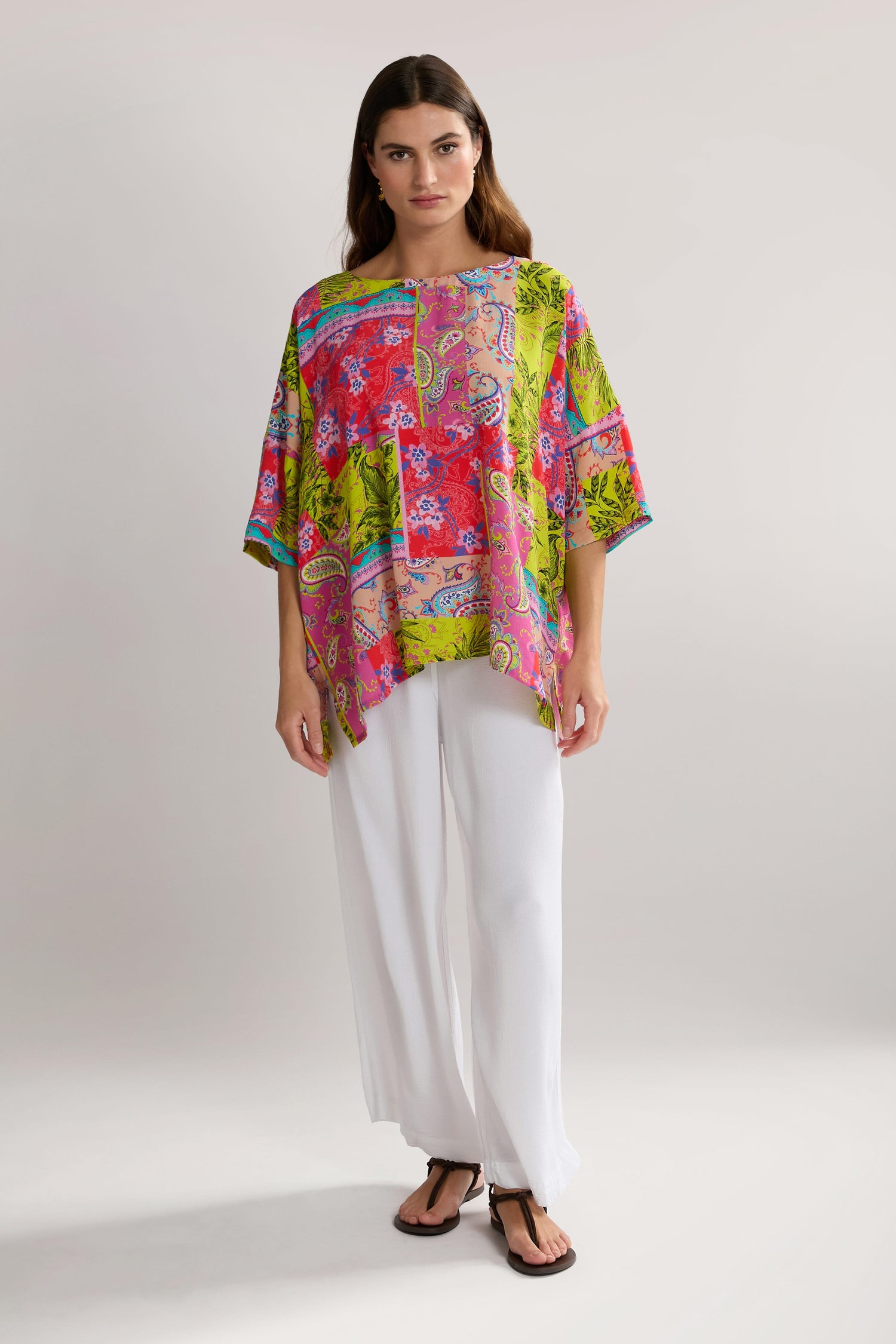 A woman stands against a plain background wearing a brightly colored, patterned Tropical Patchwork Relaxed Top made of pure viscose, paired with white pants and black sandals.