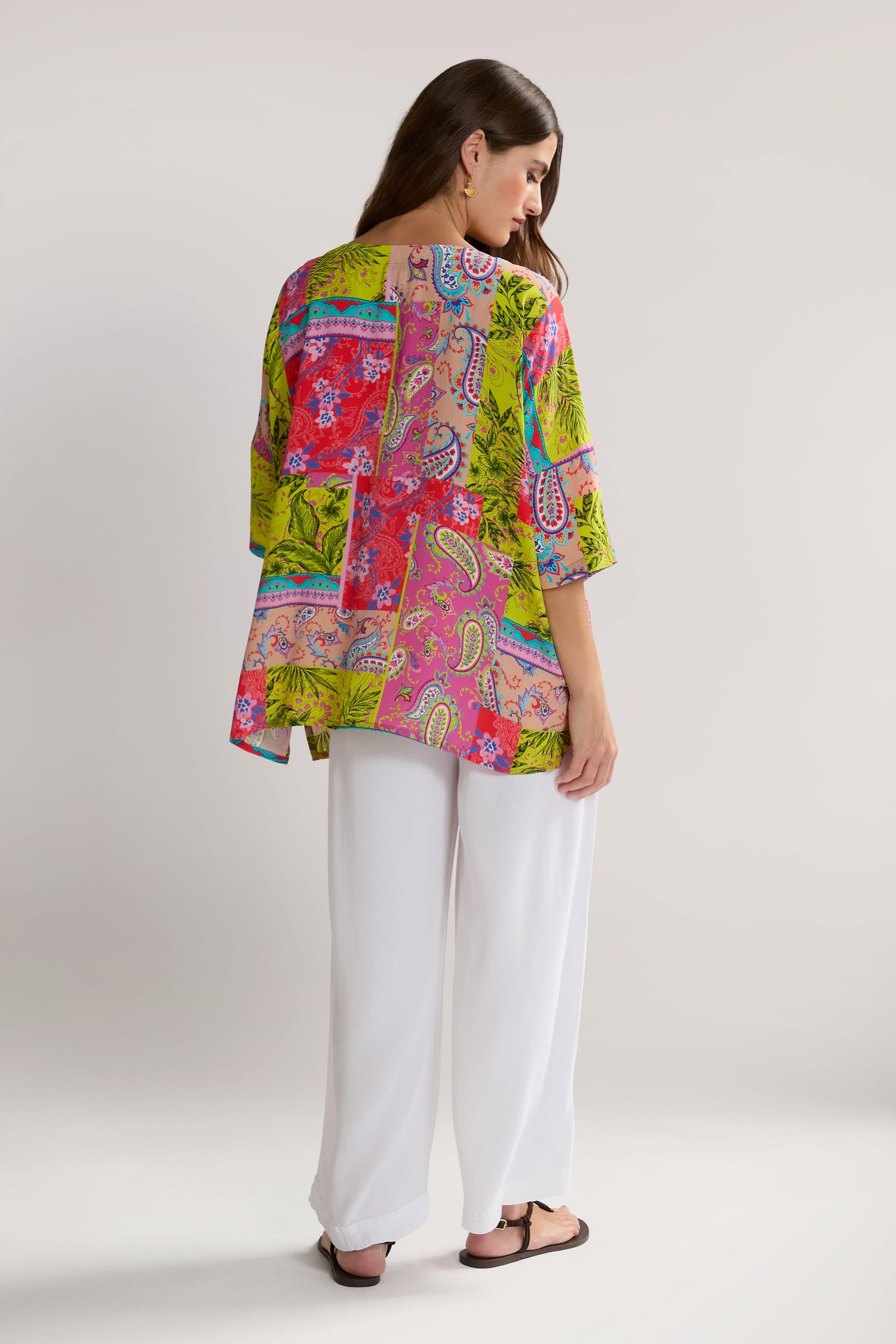 A woman with long hair stands facing away, wearing a Tropical Patchwork Relaxed Top and white pants in pure viscose. She is also wearing black sandals, embracing a bohemian style.