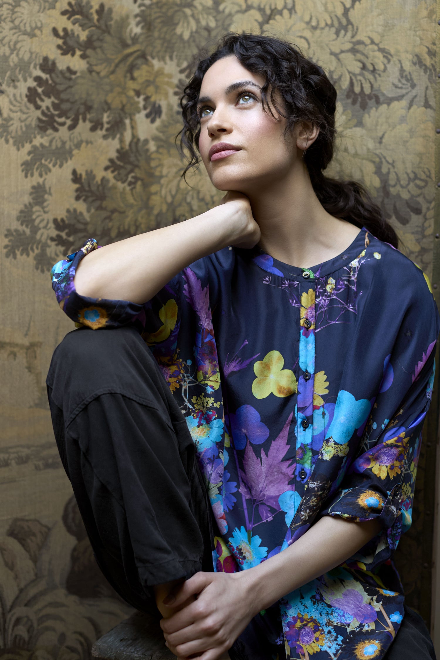 A person with curly hair and a thoughtful expression sits against a floral-patterned backdrop, donning the luxurious Blue Bouquet Silk Shirt paired with dark pants.