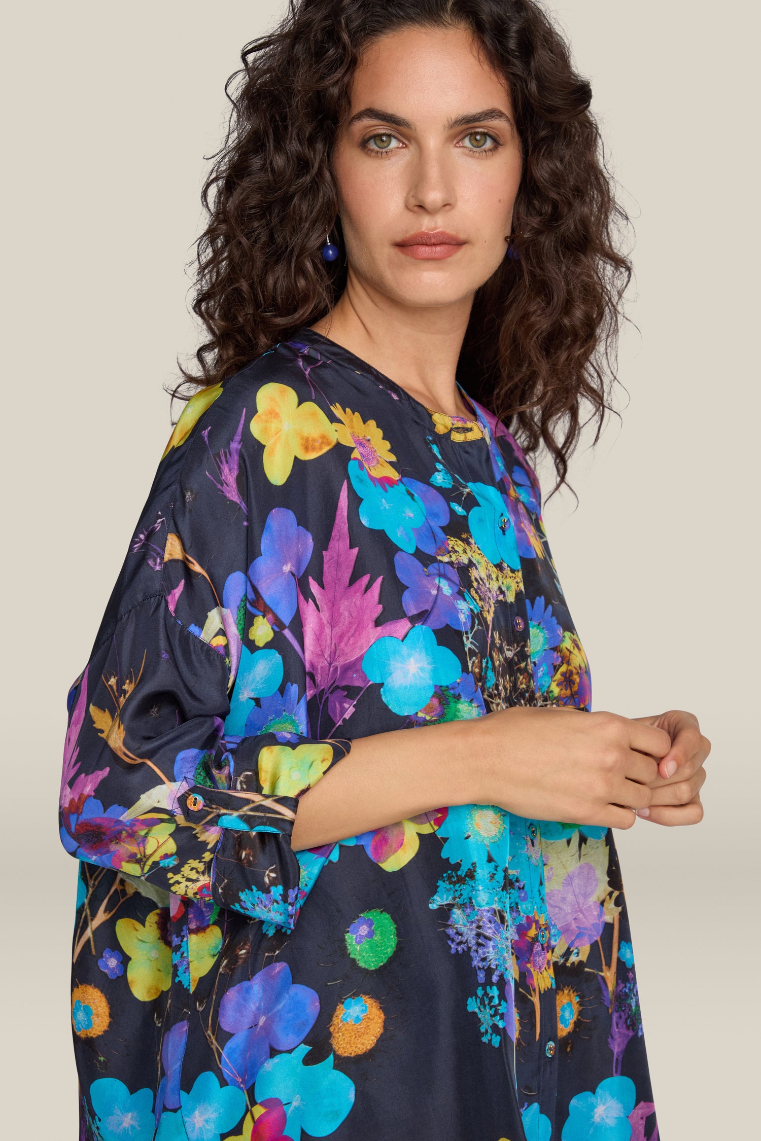 A woman with curly hair is wearing a Blue Bouquet Silk Shirt, featuring a colorful floral design and made from 100% silk. She looks at the camera with a neutral expression while holding her hands together against a plain beige background.