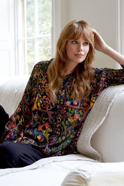 A woman sitting on a white couch wearing the Faraway Jungle Silk Boxy Shirt.