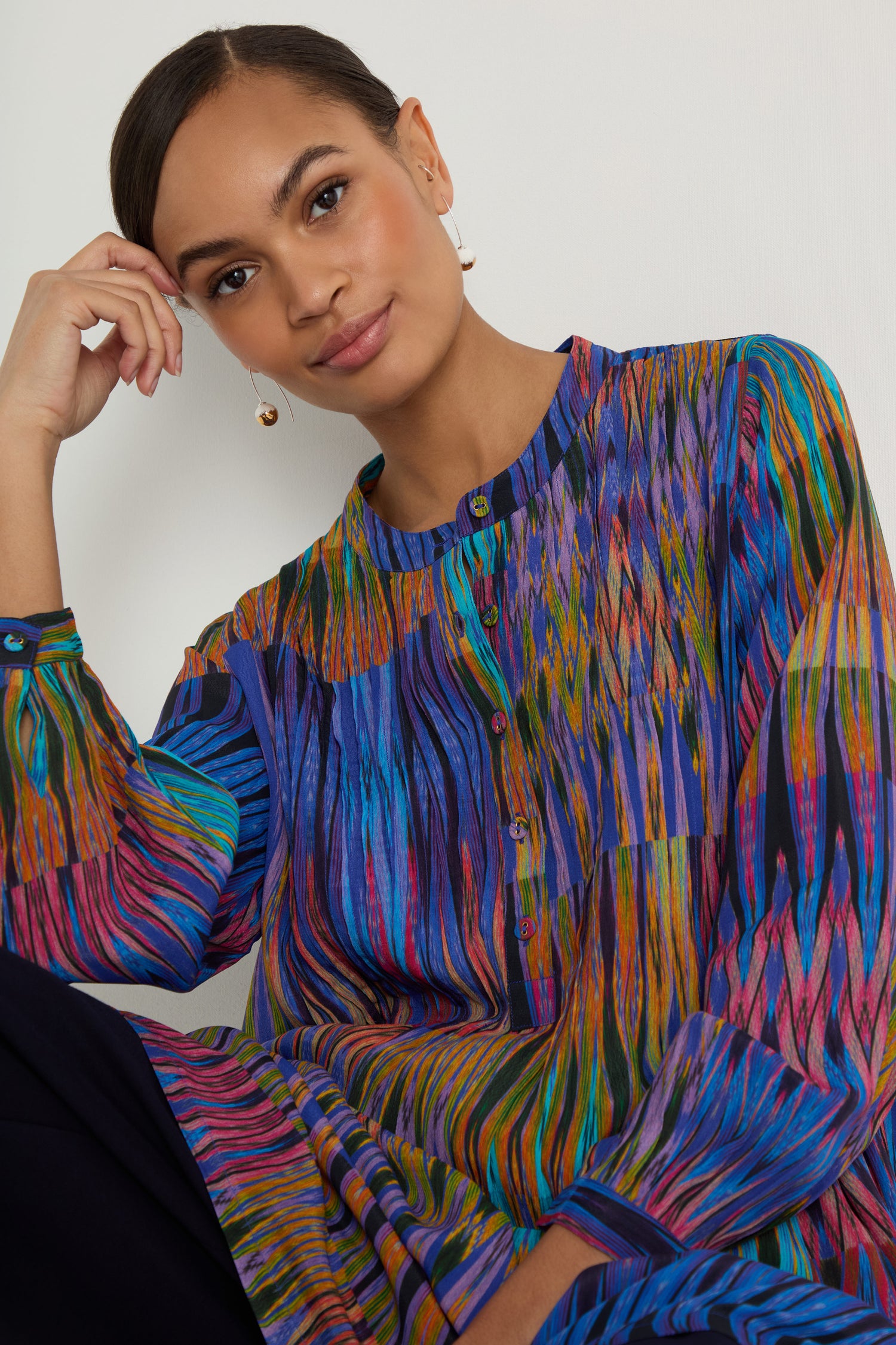 A woman with dark hair in a low bun wears the Vivid Ikat Patchwork Top with long sleeves and pearl earrings, seated against a plain light background.
