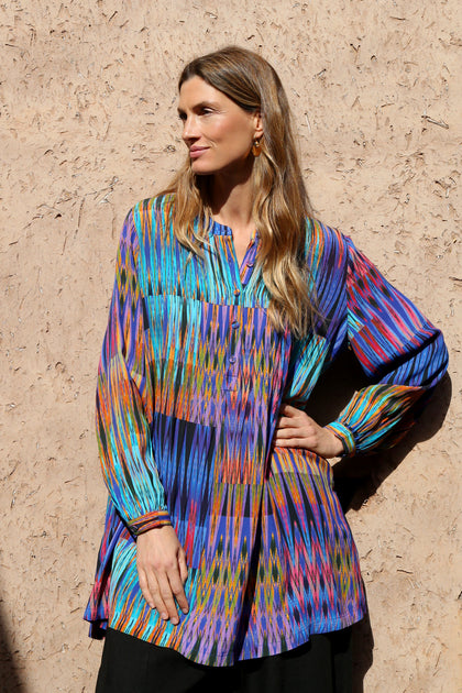 A woman with long hair stands against a textured wall, wearing the Vivid Ikat Patchwork Top and black pants, looking to the side with one hand on her hip.