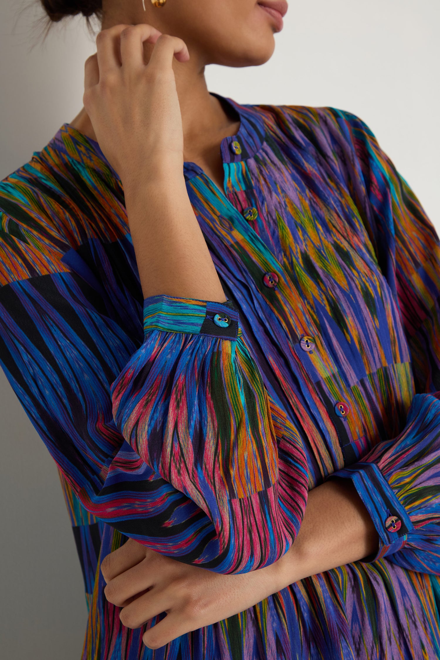 A person wears the Vivid Ikat Patchwork Top—a long-sleeved, button-up blouse with a vibrant abstract stripe pattern in blue, green, pink, and orange. The viscose top's upper buttons are casually undone.