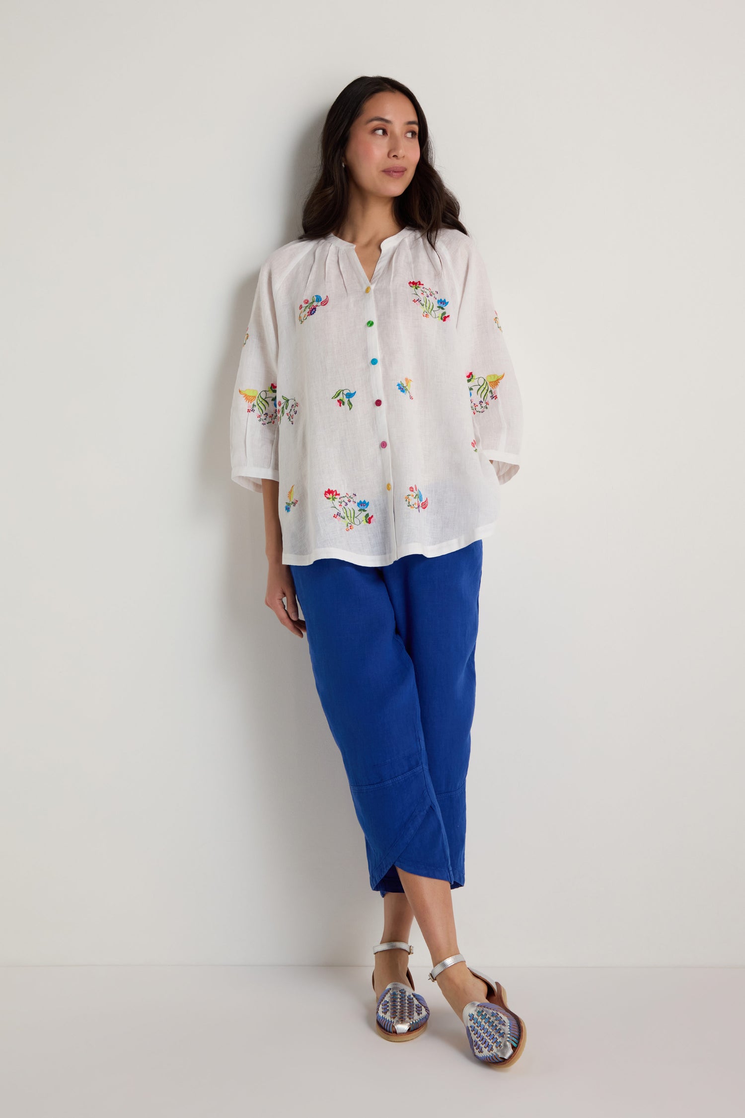 A woman stands against a white wall, wearing the Vintage Floral Embroidered Shirt, blue cropped pants, and patterned sandals.
