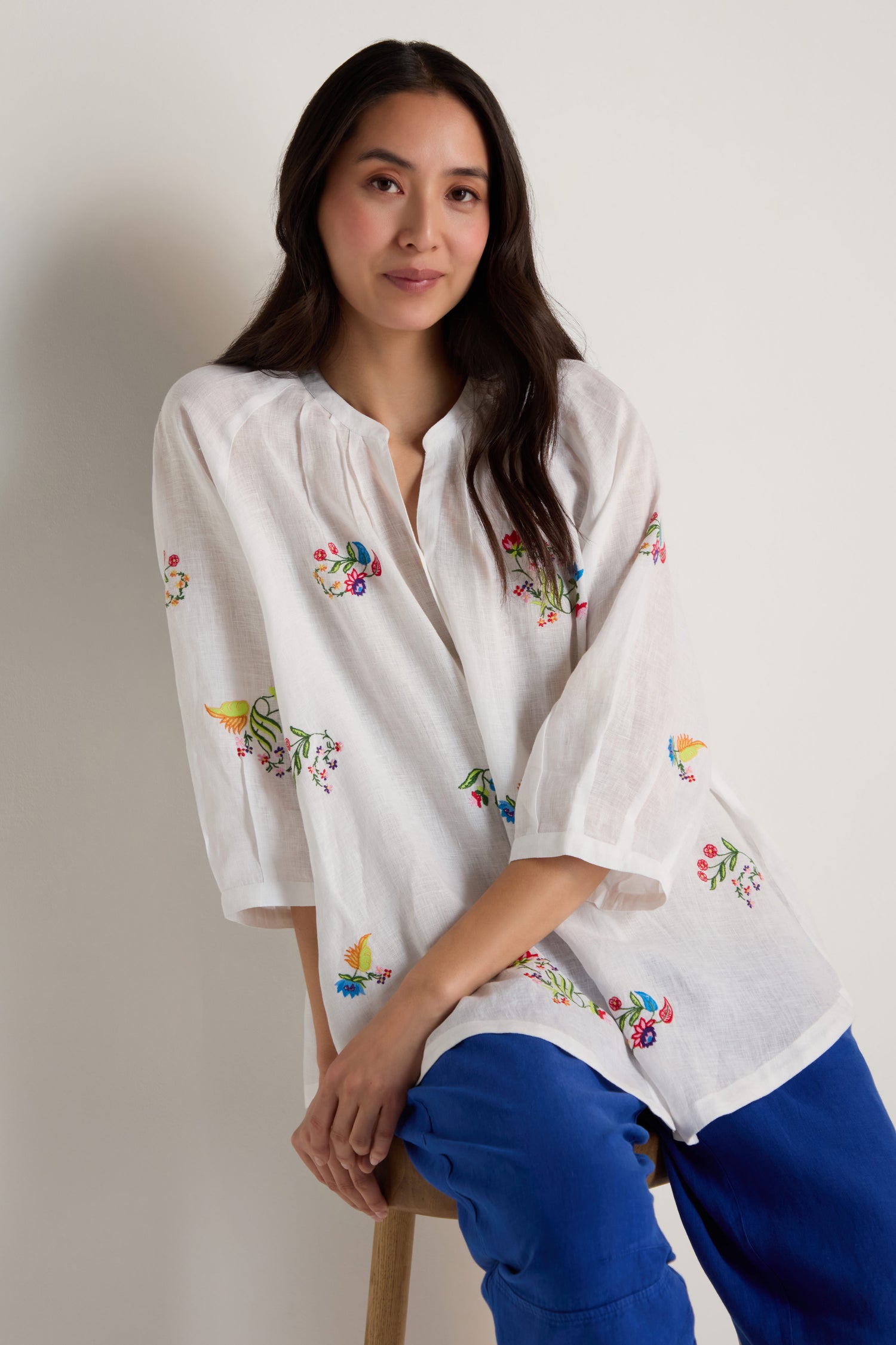 A woman in a Vintage Floral Embroidered Shirt and blue pants sits on a wooden stool against a plain background.