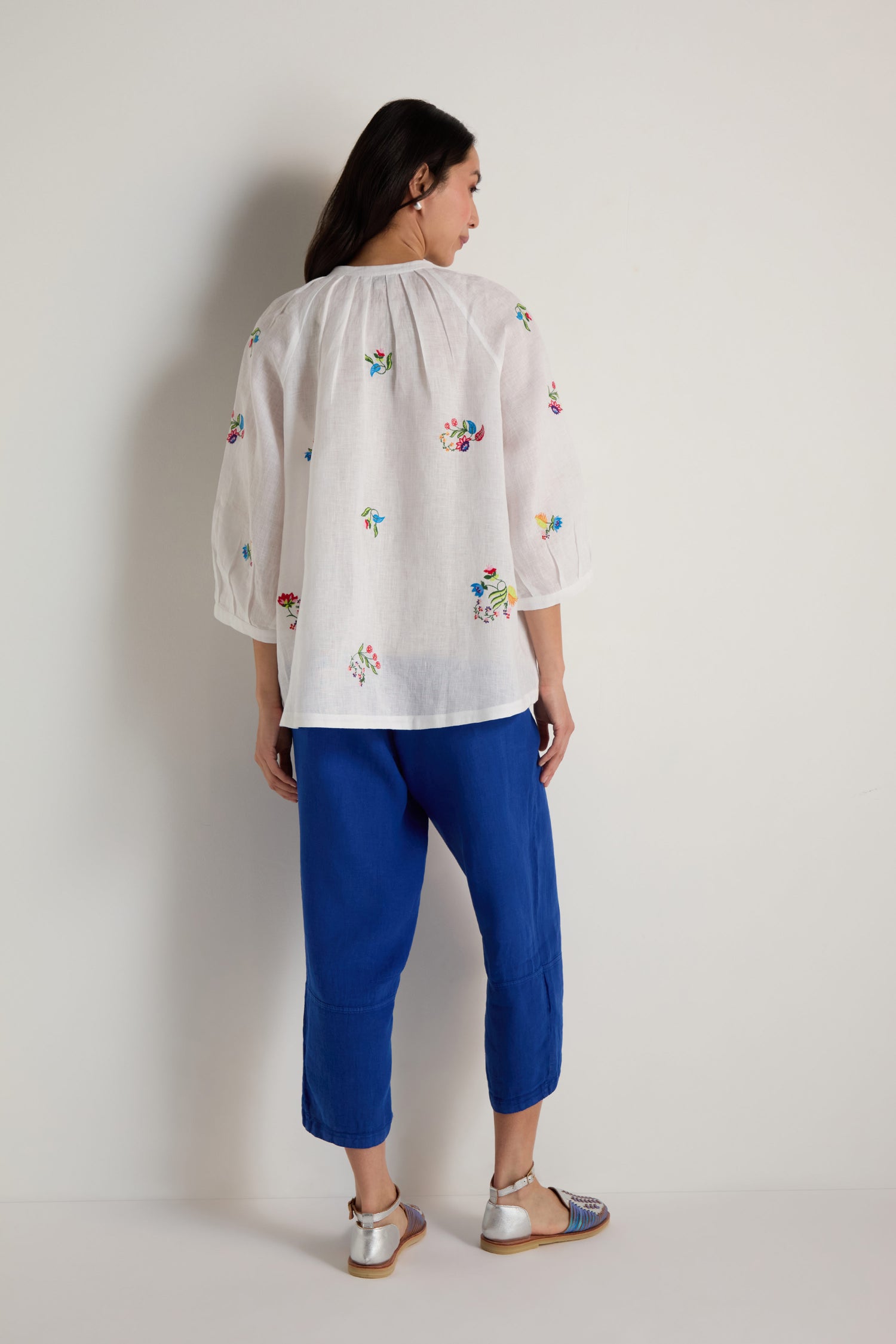 A person stands against a plain wall, facing away from the camera, wearing the Vintage Floral Embroidered Shirt, paired with blue cropped pants and silver sandals.