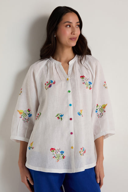 A woman wears the Vintage Floral Embroidered Shirt, featuring colorful designs and multicolored buttons, paired with blue pants, as she stands against a plain light-colored wall.