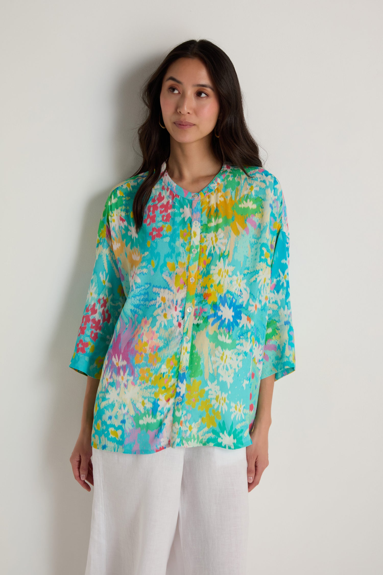 A woman stands against a plain wall wearing the Floral Oasis Boxy Shirt, a turquoise floral viscose crepe top with white, yellow, and pink accents, paired with white pants.
