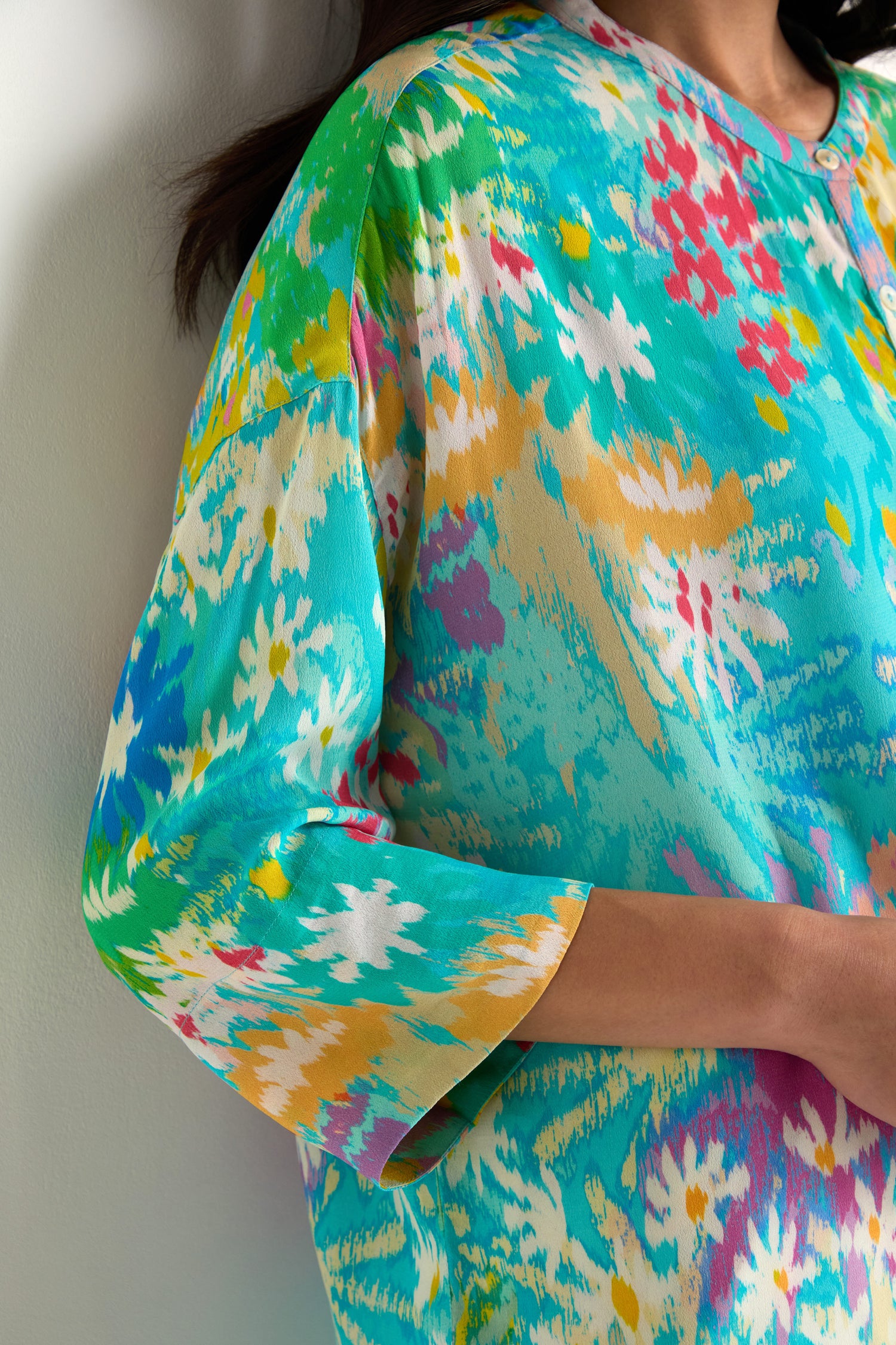 A person wearing the Floral Oasis Boxy Shirt, an oversized, brightly colored floral top with turquoise, yellow, white, and pink accents, stands against a light-colored wall.