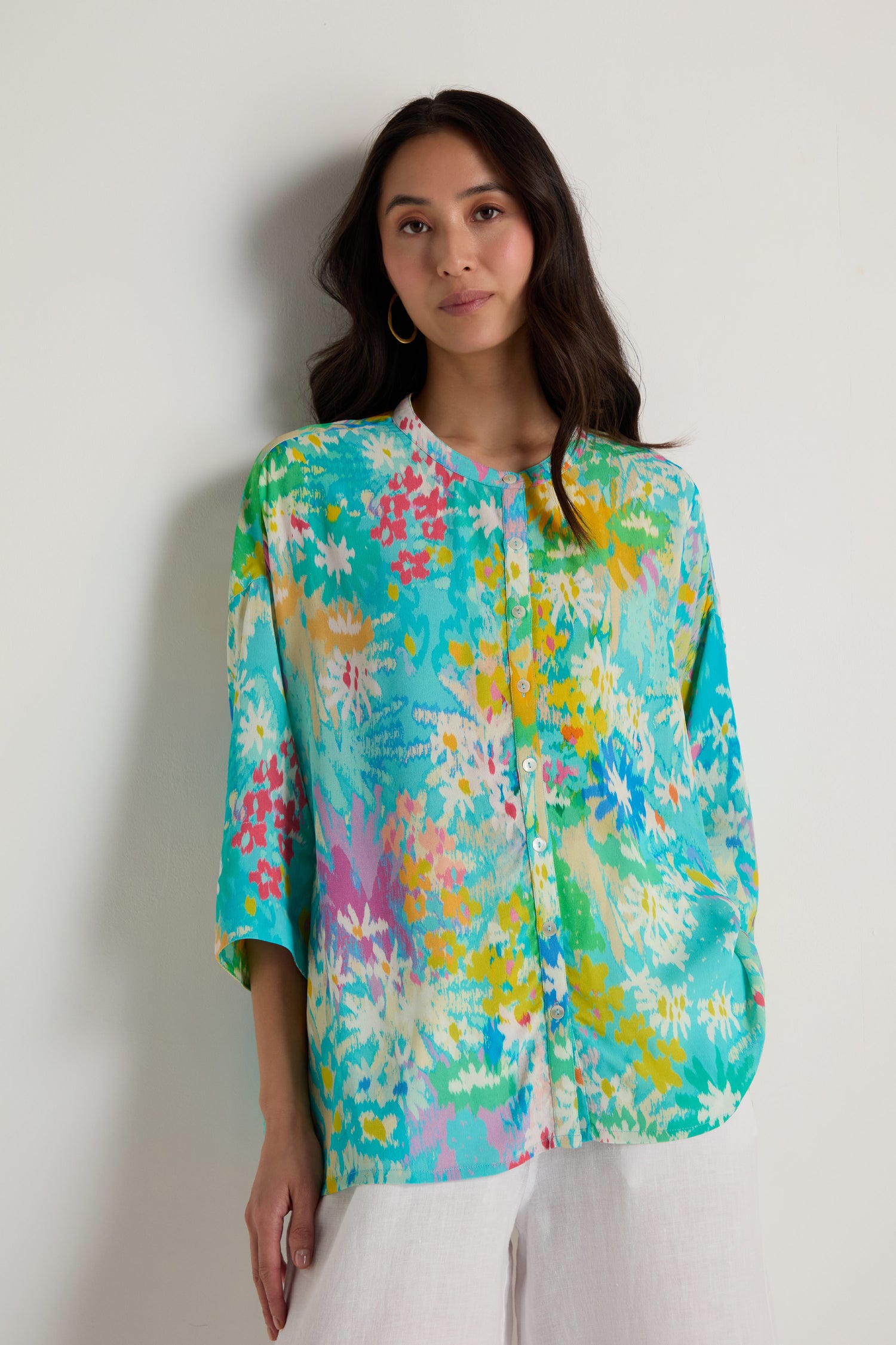 A woman stands against a plain wall wearing the Floral Oasis Boxy Shirt, featuring a brightly colored floral design in an oversized fit, paired with white pants.
