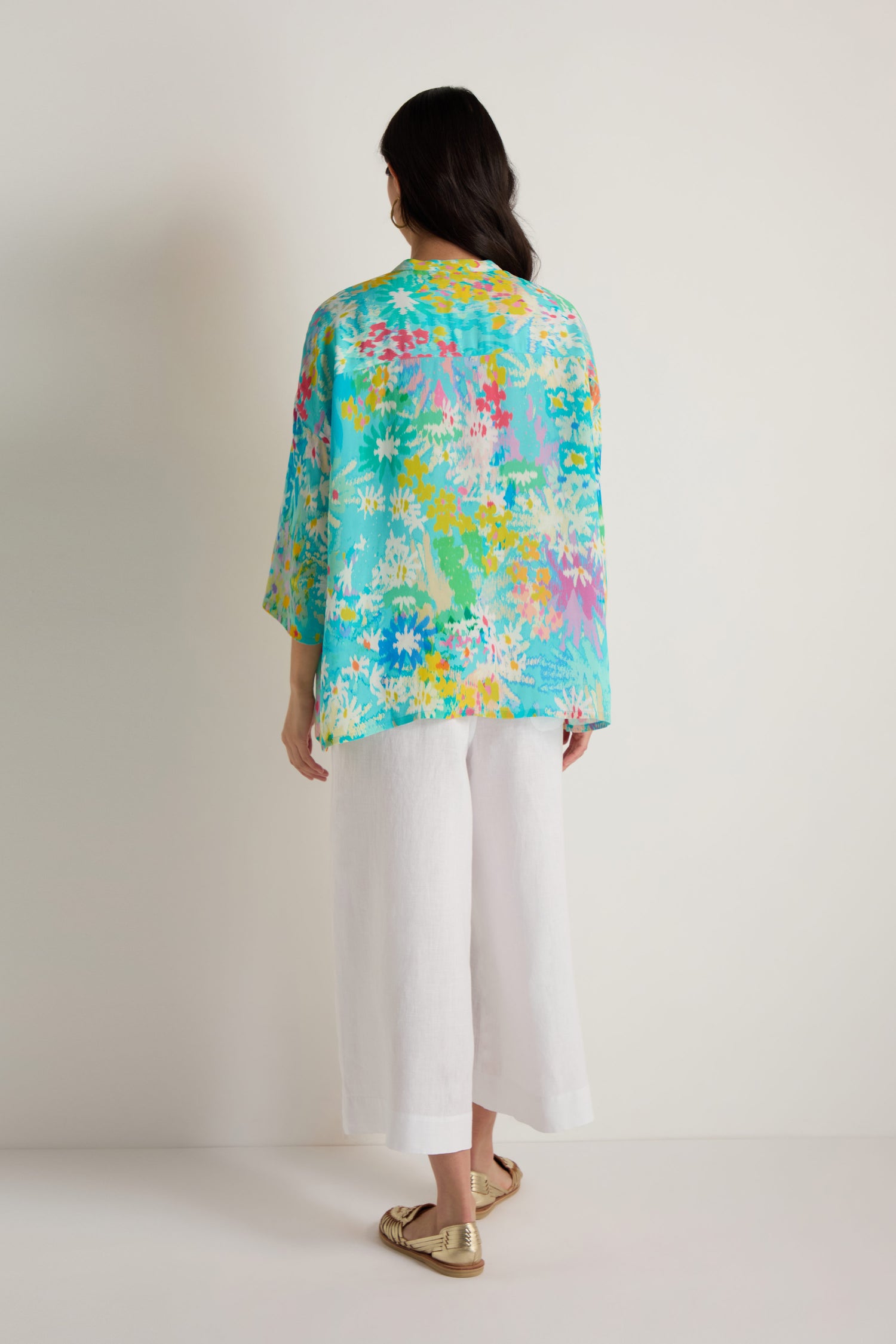 A woman faces away, wearing the Floral Oasis Boxy Shirt in turquoise viscose crepe, paired with white wide-leg pants and gold flats against a plain light background.