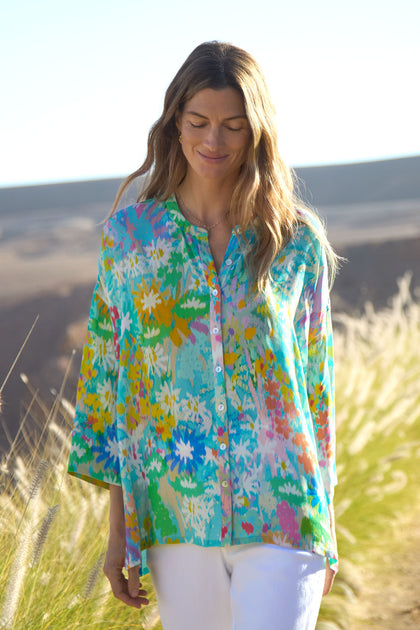 A woman walks outdoors in the Floral Oasis Boxy Shirt and white pants, with tall grass and hills in the background.