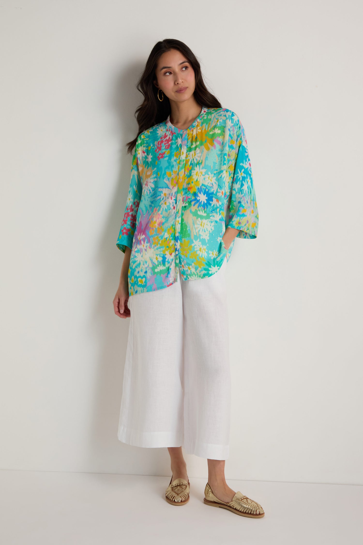 A woman stands against a plain white background wearing the Floral Oasis Boxy Shirt in turquoise floral viscose crepe with an oversized fit, paired with white wide-leg pants and beige woven flats.
