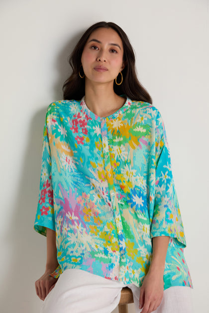 A woman in the Floral Oasis Boxy Shirt, featuring a colorful floral print and oversized fit, pairs it with white pants while sitting on a stool against a plain white wall.