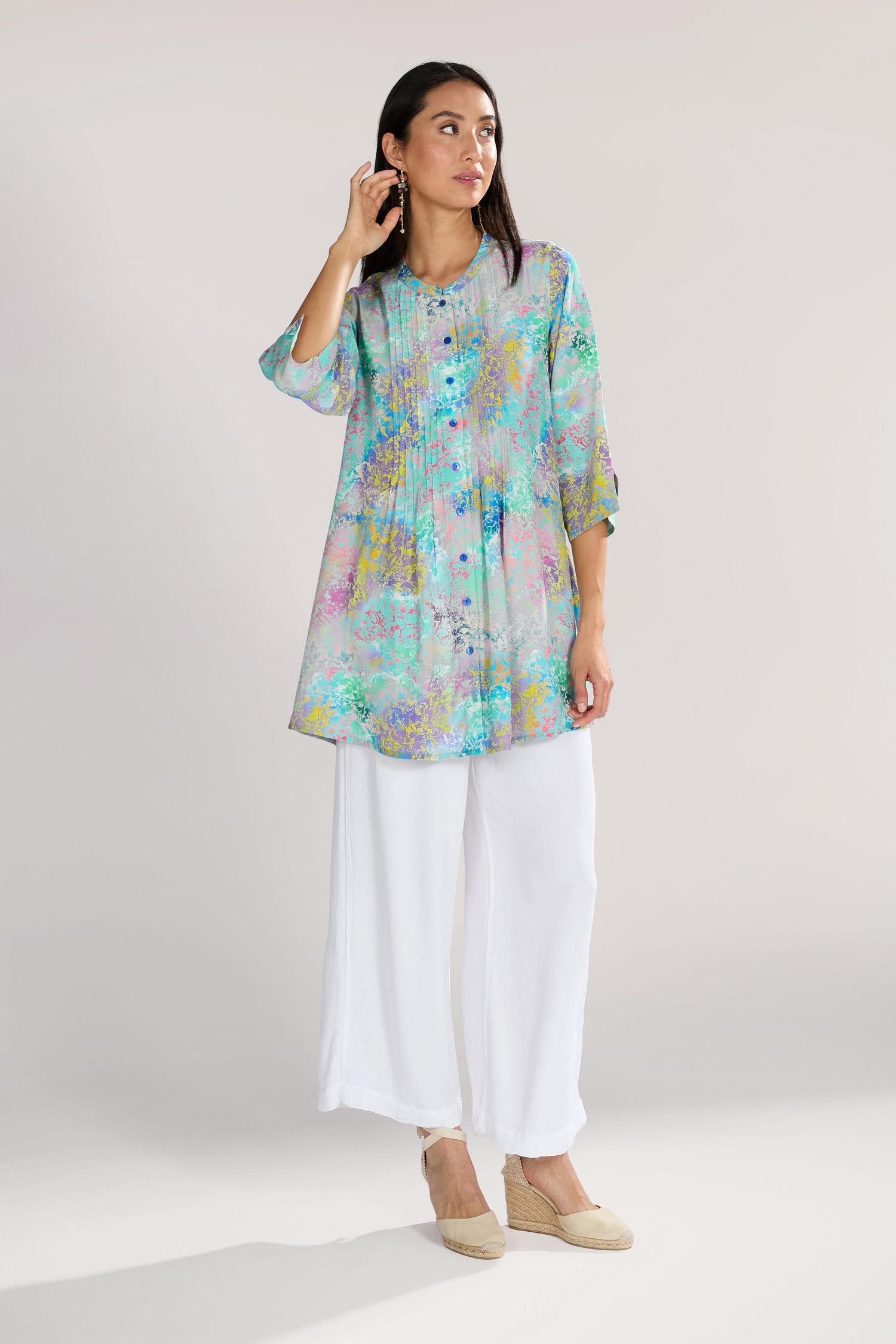 A woman in a colorful, long-sleeved tunic and white pants stands against a neutral background, touching her ear with one hand. Her flowing viscose fabric hints at the elegance of the Marbled Cloud Pleat Shirt she wears.