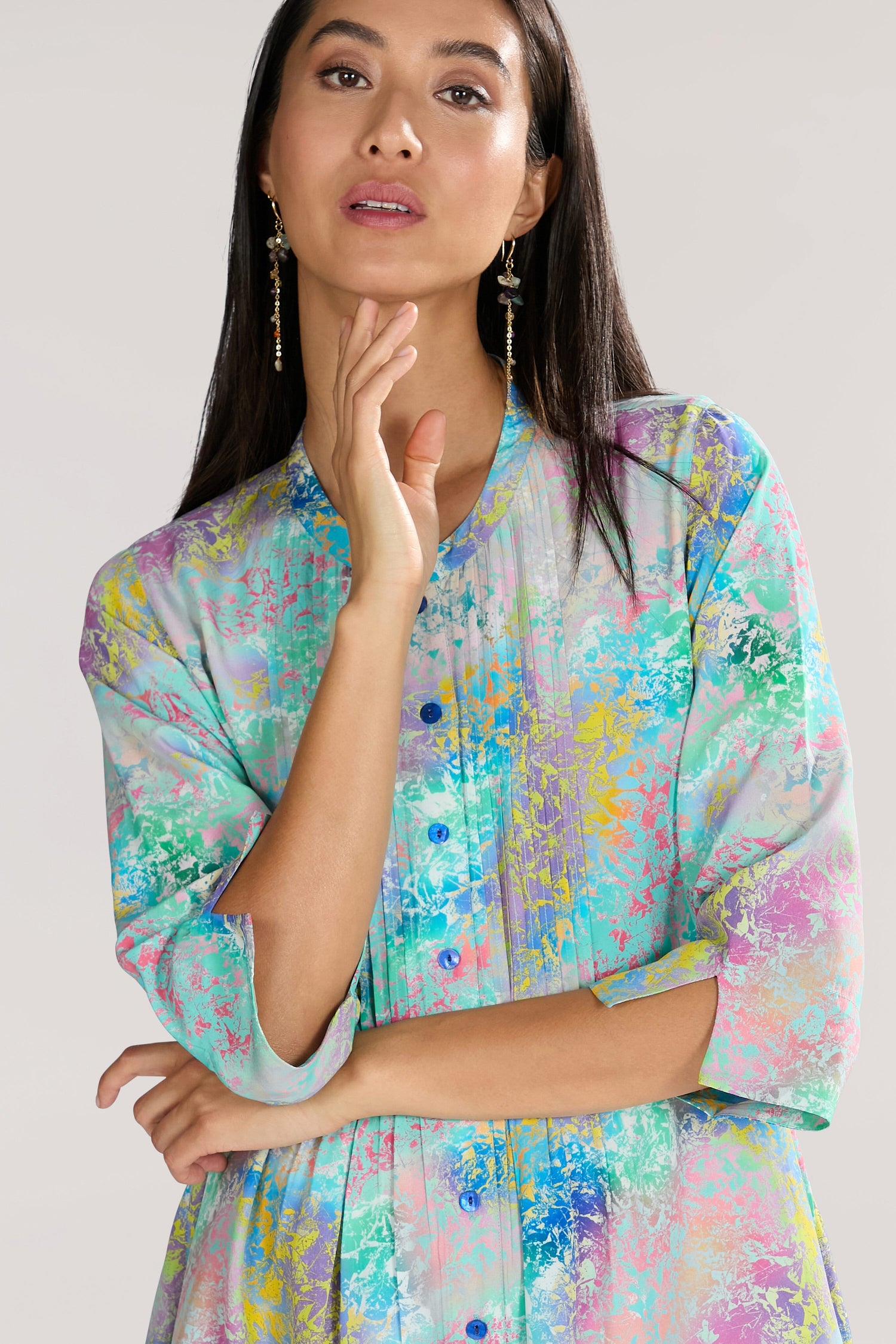 A woman wearing an elegant **Marbled Cloud Pleat Shirt** stands with her right hand touching her face and the left arm crossed. She has long dark hair and is gazing forward with a neutral expression.