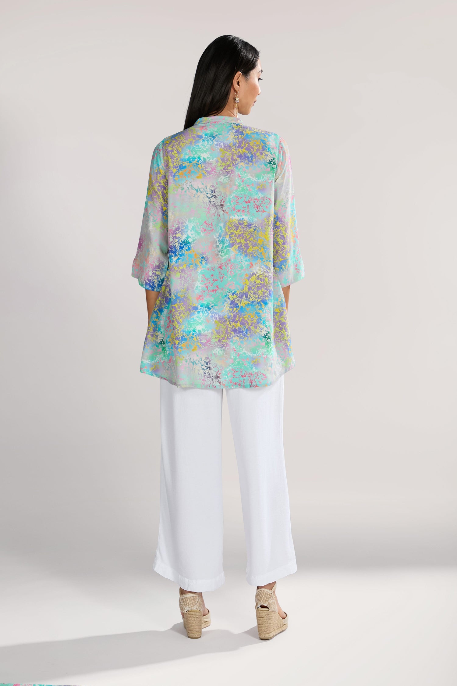 A person stands facing away from the camera, wearing an elegant Marbled Cloud Pleat Shirt with shades of blue, purple, and green and white pants. They have long dark hair, are donning wedge sandals, and the flowing viscose fabric creates a breezy silhouette.