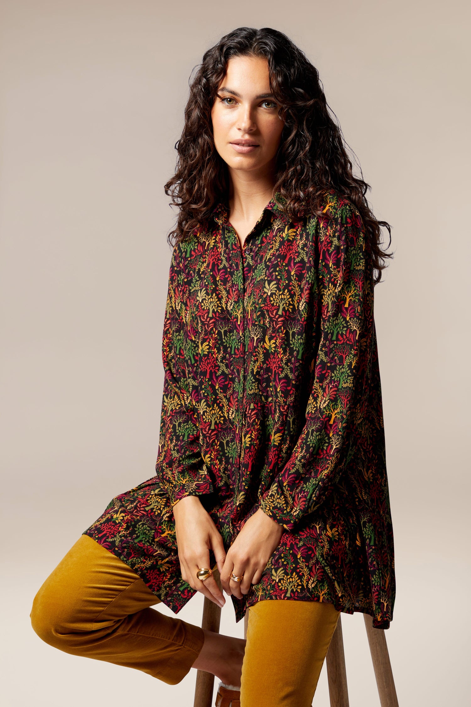 A woman is sitting on a stool wearing a Magical Forest Print Shirt in autumnal tones.