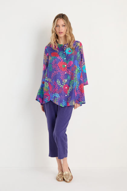 A woman stands against a white wall, dressed in the Bright Blooming Garden Silk Shirt with a botanical floral print, paired with purple cropped pants and gold flat shoes.