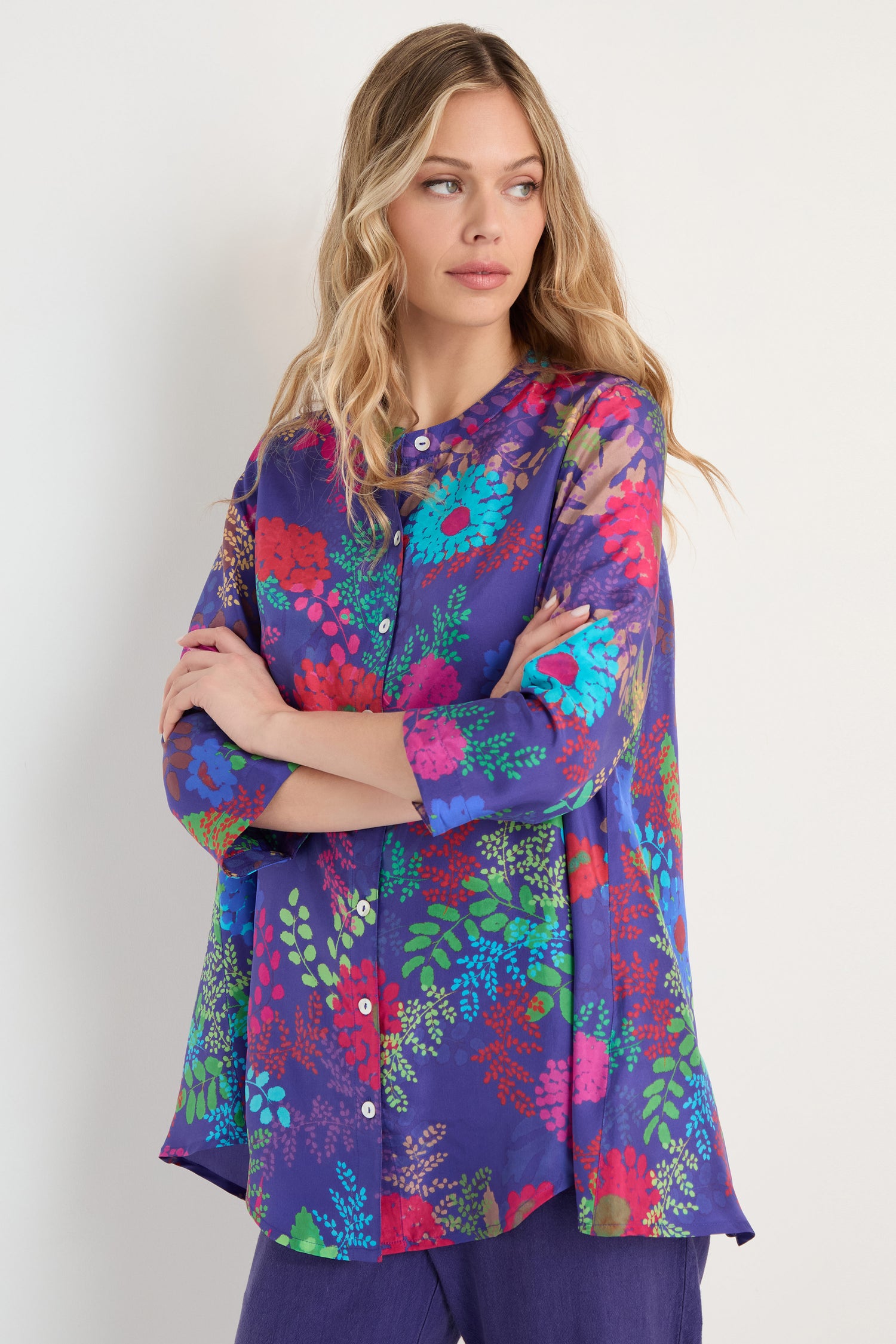 A woman in the Bright Blooming Garden Silk Shirt with long sleeves and a botanical floral print pairs it with purple pants, standing against a plain light background.