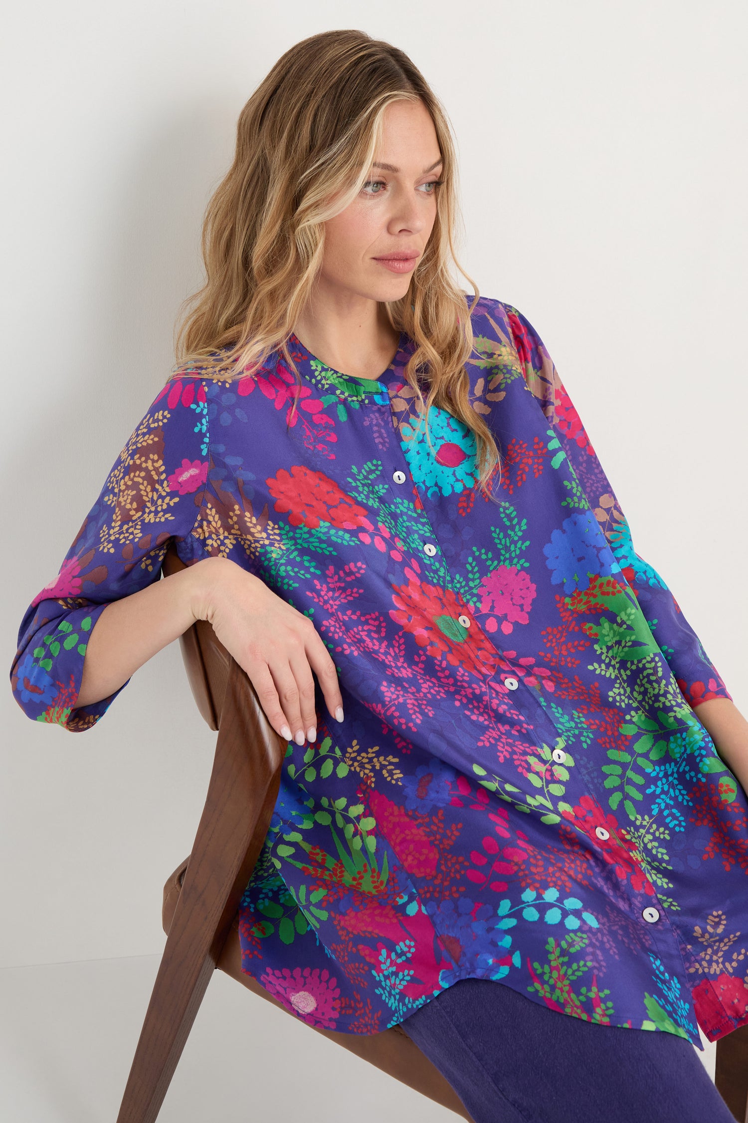 A woman with wavy blonde hair sits on a wooden chair, wearing the Bright Blooming Garden Silk Shirt with a colorful floral print and dark pants against a plain light background.
