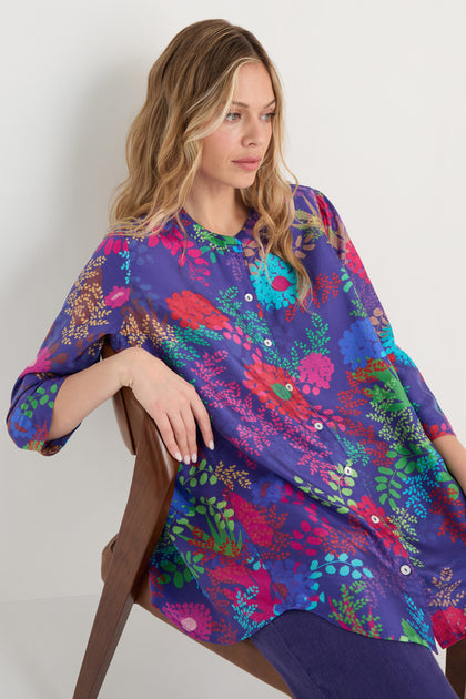 A woman with wavy blonde hair sits on a wooden chair, wearing the Bright Blooming Garden Silk Shirt with a colorful floral print and dark pants against a plain light background.