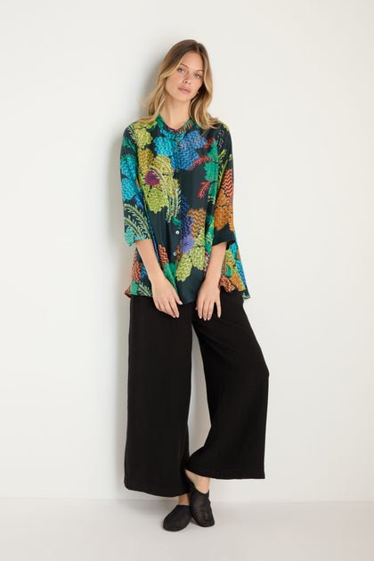 A woman stands against a plain wall, wearing the Pixelated Petals Print Silk Shirt with wide sleeves, paired with black wide-leg pants and black shoes.