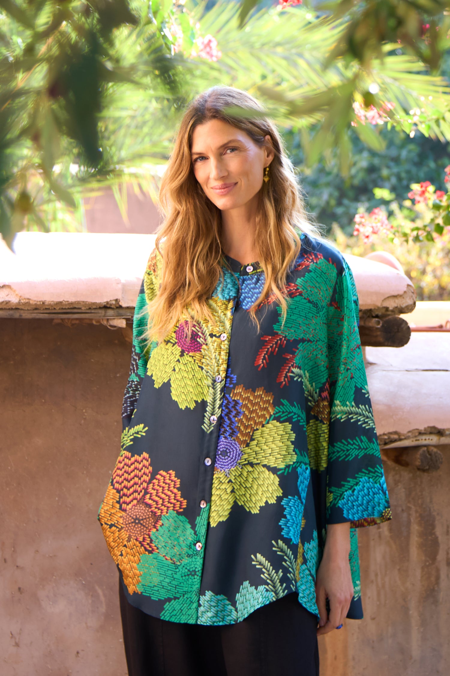 A woman stands outdoors against greenery and a stone wall, wearing the Pixelated Petals Print Silk Shirt—a loose, colorful silk top featuring a vibrant floral print—paired with black pants.