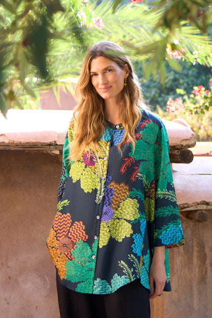 A woman stands outdoors against greenery and a stone wall, wearing the Pixelated Petals Print Silk Shirt—a loose, colorful silk top featuring a vibrant floral print—paired with black pants.