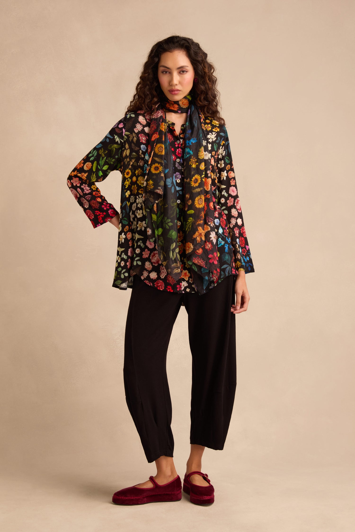 A woman stands against a beige background, dressed in the Midnight Garden Viscose Shirt with a flared silhouette, paired with a matching floral scarf, loose black pants, and red slippers.