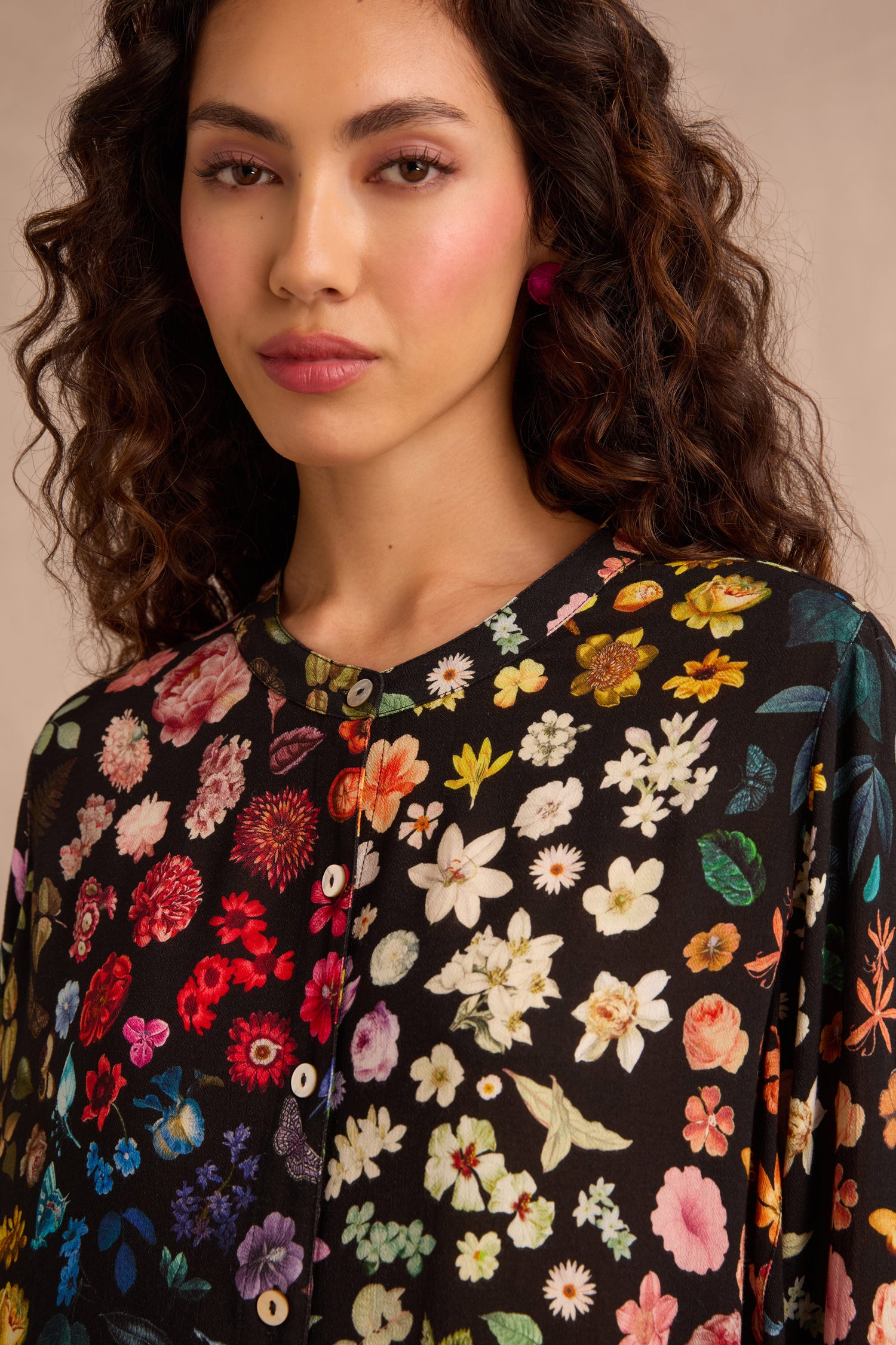 A woman with wavy hair models the Midnight Garden Viscose Shirt—a black button-up adorned with a vibrant floral print and a softly flared silhouette—while standing against a neutral background.