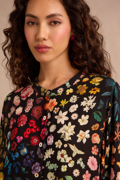 A woman with wavy hair models the Midnight Garden Viscose Shirt—a black button-up adorned with a vibrant floral print and a softly flared silhouette—while standing against a neutral background.