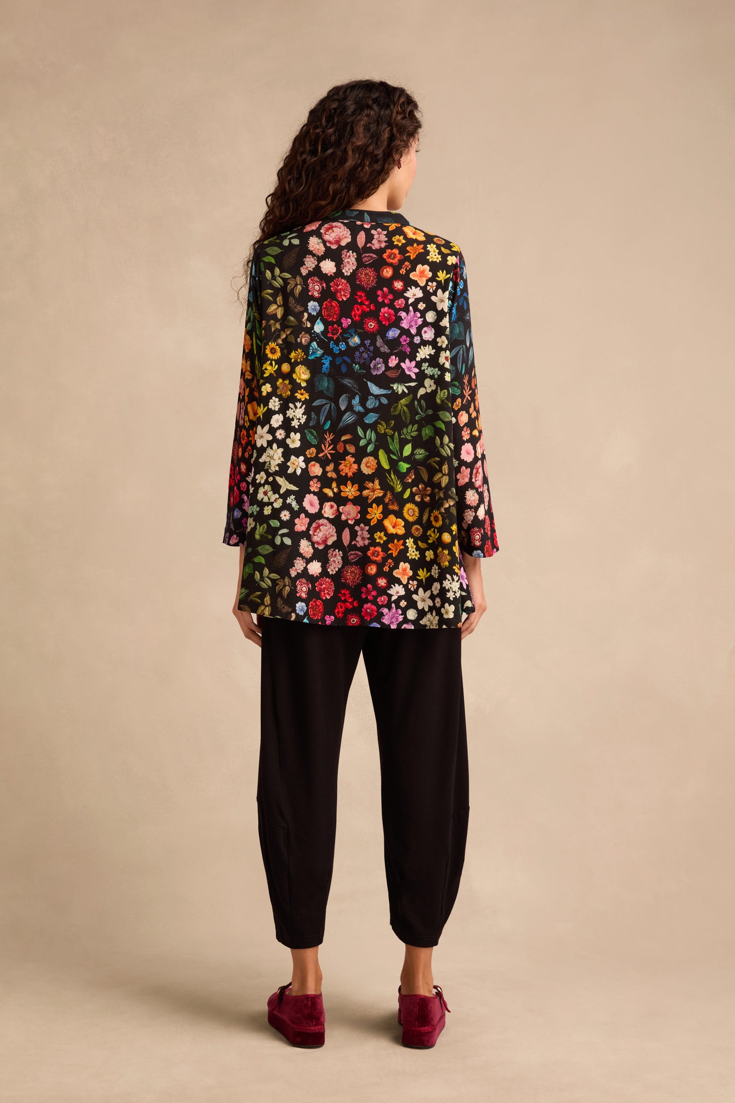 A person stands facing away, showcasing the Midnight Garden Viscose Shirt—known for its vibrant floral pattern and flared silhouette—paired with black pants and red shoes. Their curly hair is visible from behind, set against a beige background.