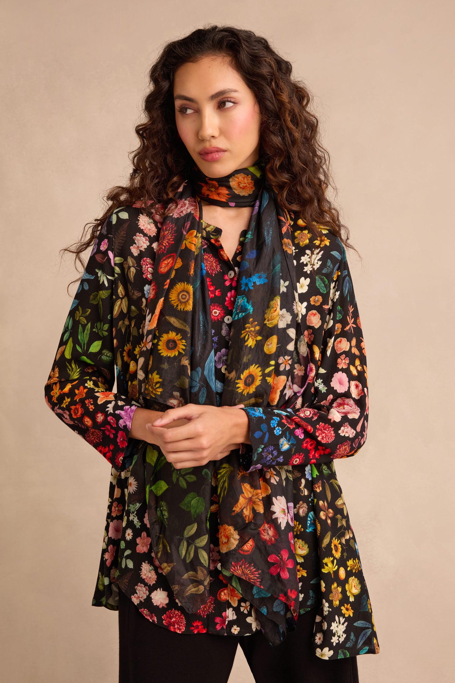 A woman with long curly hair models the Midnight Garden Viscose Shirt, paired with a matching scarf. She stands against a plain beige background, her hands clasped in front, while the shirt’s flared silhouette lends elegance to her look.