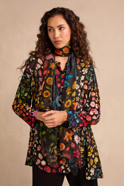 A woman with long curly hair models the Midnight Garden Viscose Shirt, paired with a matching scarf. She stands against a plain beige background, her hands clasped in front, while the shirt’s flared silhouette lends elegance to her look.