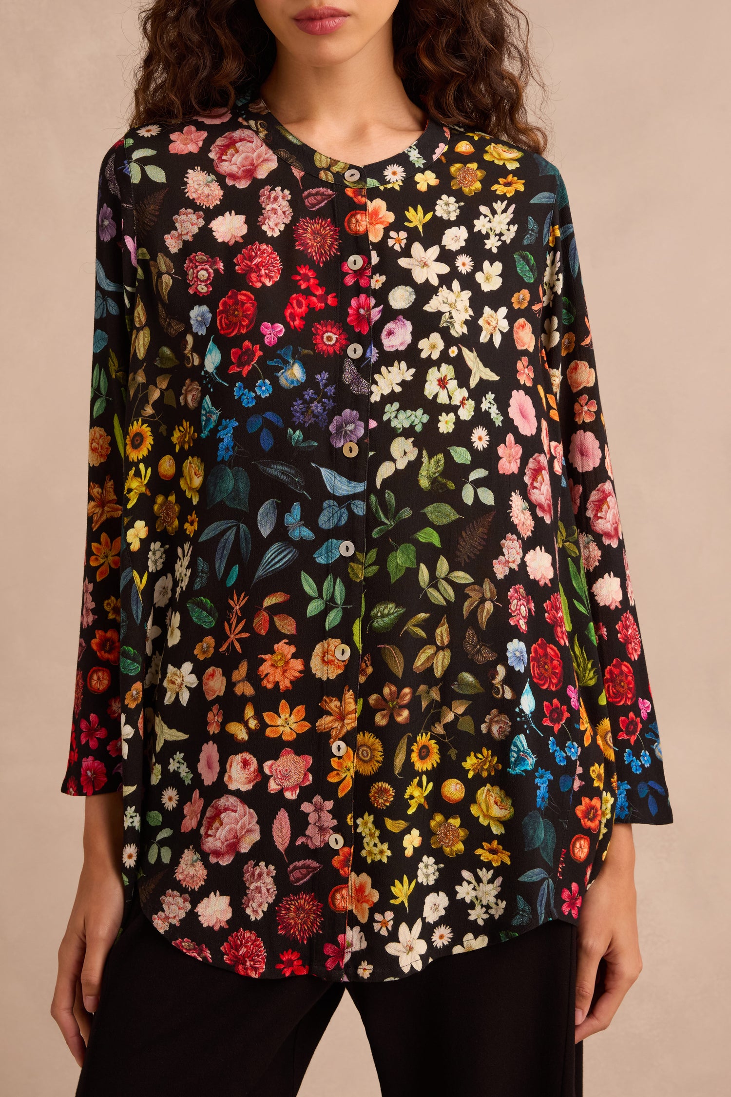 A woman stands against a neutral background, wearing the Midnight Garden Viscose Shirt—a black long-sleeve blouse featuring a colorful floral print and a subtly flared silhouette.