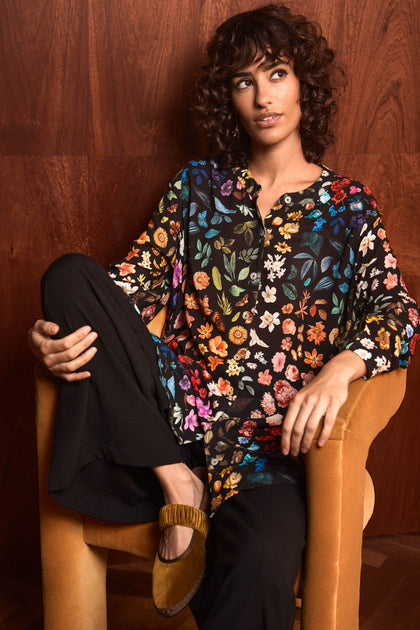 A person with curly hair sits in a tan armchair, wearing the Midnight Garden Viscose Shirt, black pants, and brown flat shoes, against a wooden wall.
